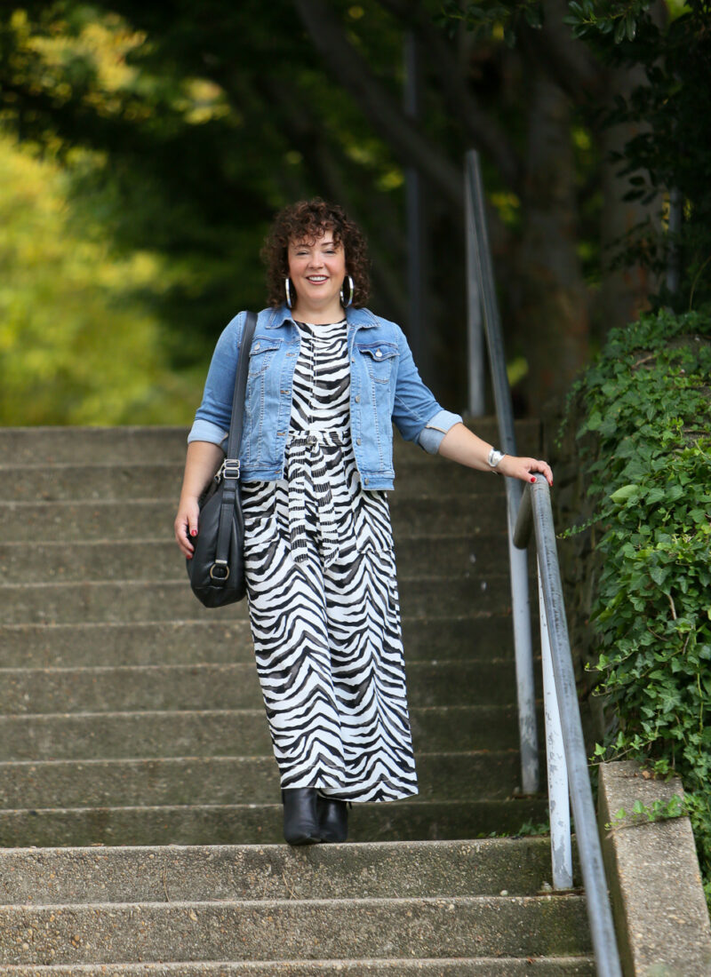 Styling a summer jumpsuit for fall - tips on how to do this successfully by Wardrobe Oxygen who is wearing a zebra print Banana Republic jumpsuit with a denim jacket and black ankle booties by Clark's and carrying a Rough & Tumble handbag in black leather.