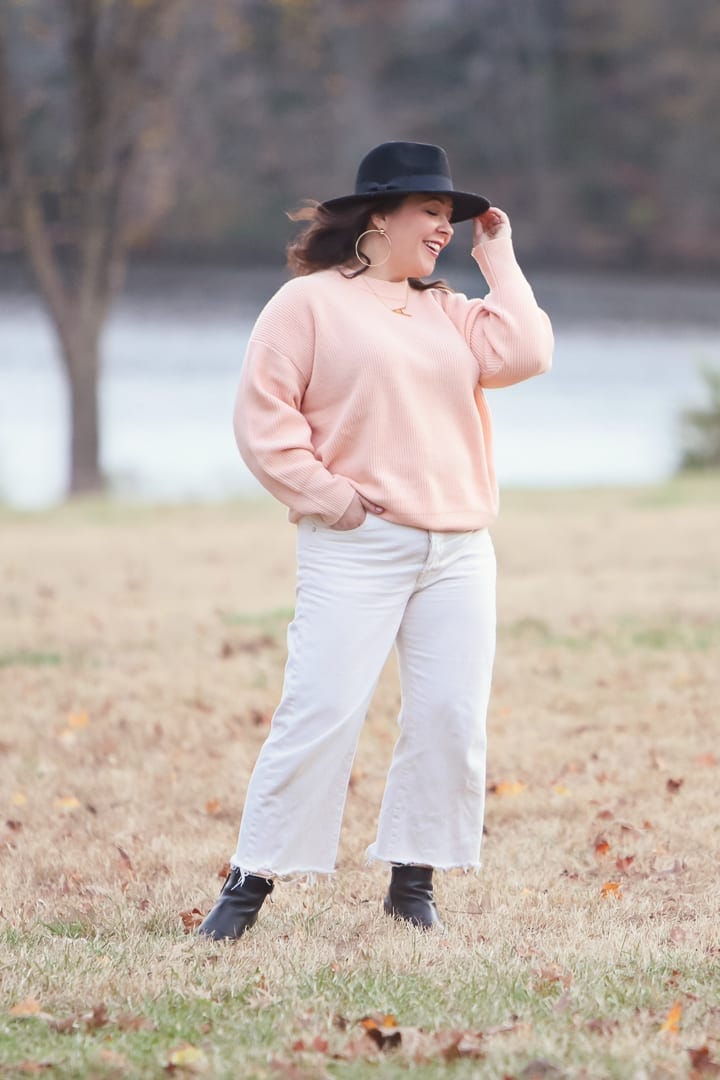 woman in peach sweater and white wide leg cropped jeans standing in a field tipping her hat