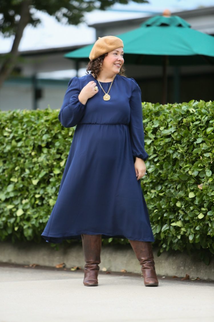 woman in a navy long sleeve dress with a camel beret, gold pendant necklace, and brown knee high boots
