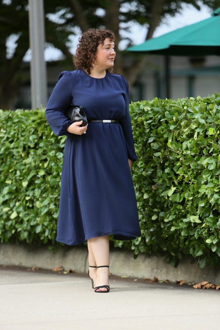 woman in a navy dress with a black leather skinny belt with rhinestone buckle, black leather clutch purse, and black strappy heels