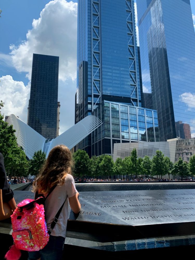 young girl looking into the 9/11 memorial fountain with the World Trade Center buildings and the Oculus in the distance.