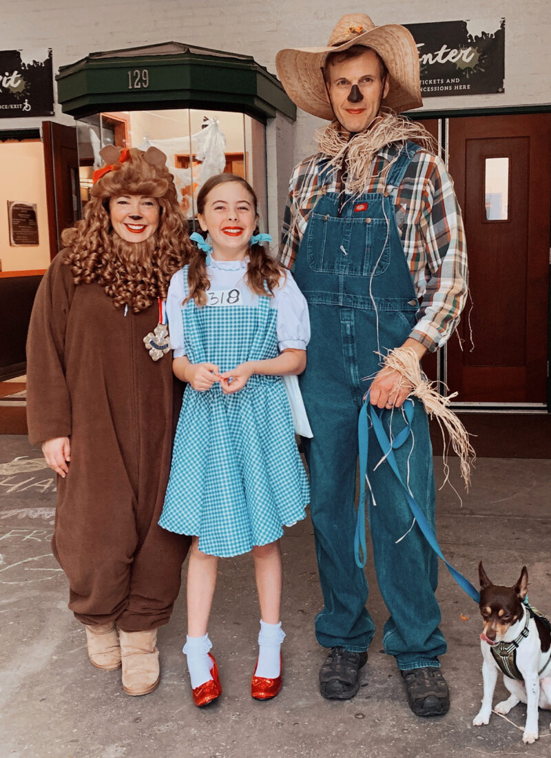 a woman dressed as the cowardly lion, young girl dressed as Dorothy Gale, man dressed as a scarecrow holding onto the leash of a small dog