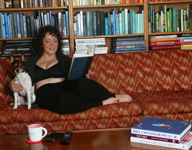 woman reading a book on a couch with her dog. She is wearing a wireless bra, black leggings, and a gray cardigan.