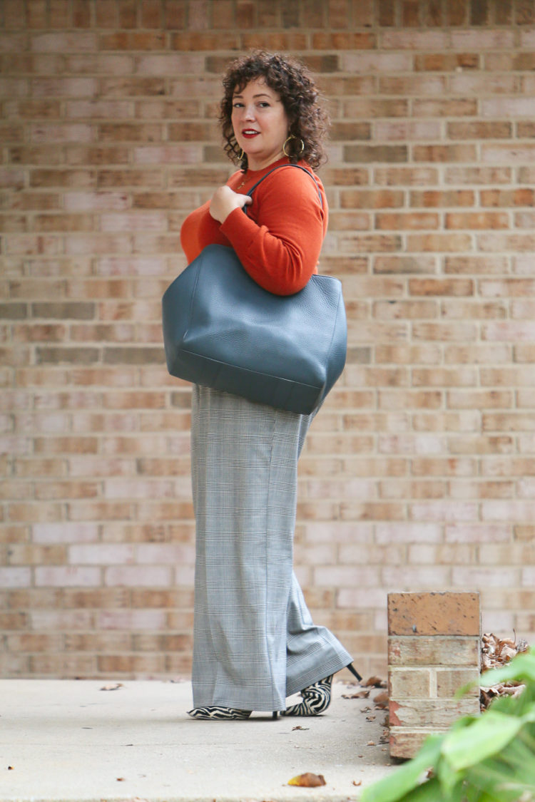 woman in an orange sweater and gray plaid pants carrying a teal blue leather tote walking out of an office building