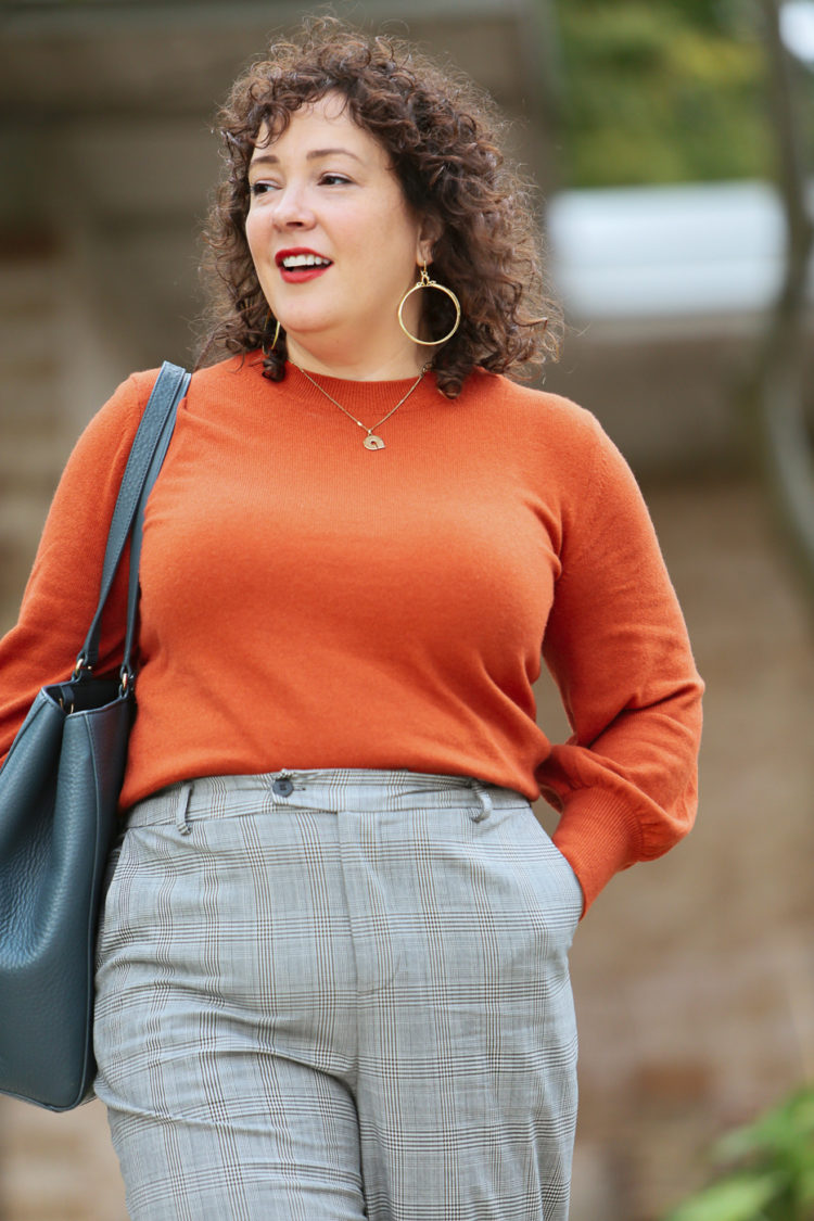 woman in an orange sweater and gray plaid pants carrying a teal blue leather tote walking out of an office building