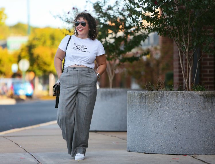 woman walking along a strip mall in white graphic t-shirt tied at her waist with gray glen plaid wide leg trousers