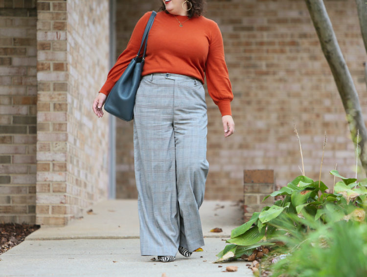 woman in an orange long sleeved sweater tucked into gray plaid pants walking o a sidewalk looking to the side