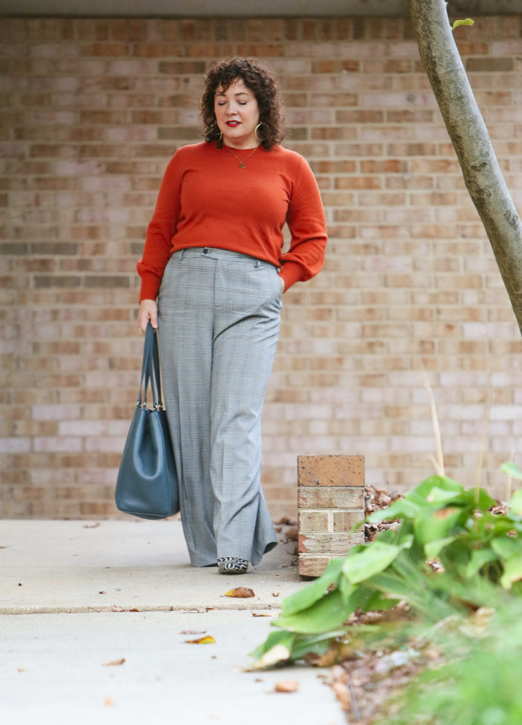 woman in an orange sweater and plaid pants walking down a sidewalk