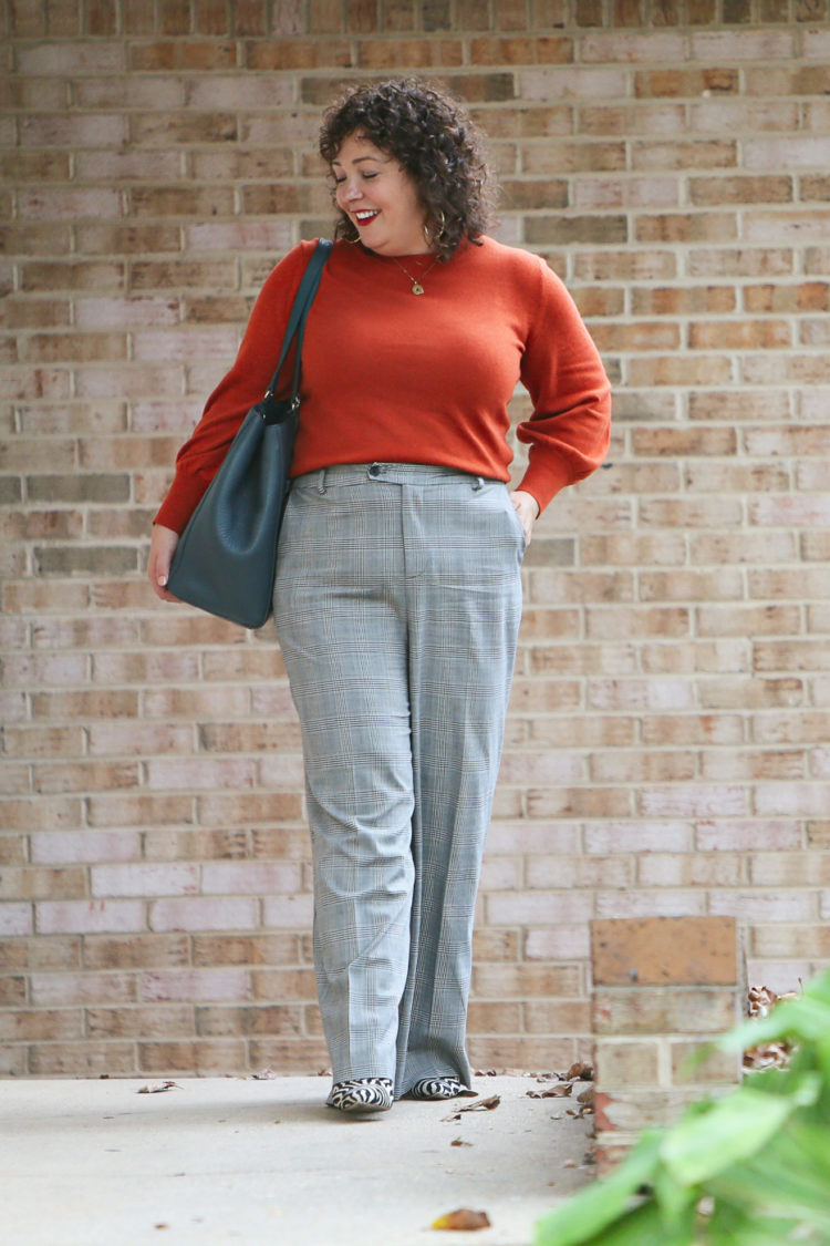 woman in an orange sweater and gray plaid pants carrying a teal blue leather tote walking out of an office building