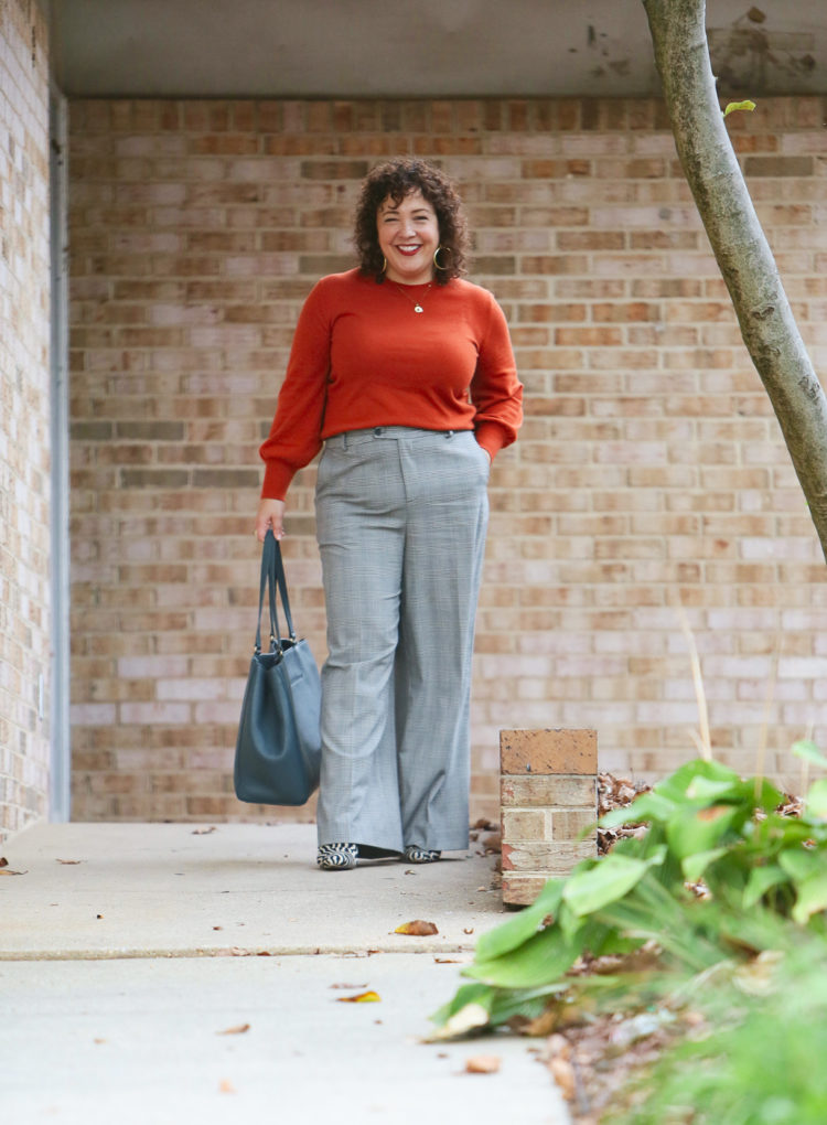 woman in an orange sweater and gray plaid pants carrying a teal blue leather tote walking out of an office building