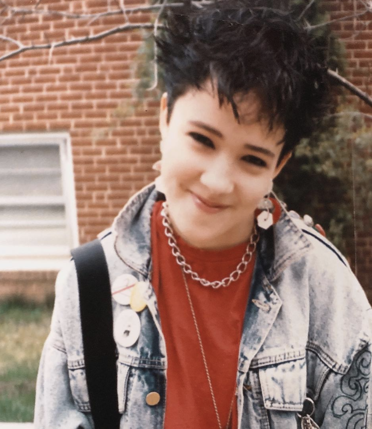 Photo of a 9th grade girl with spiky black hair, a lot of black eyeliner, an acid washed denim jacket covered with pins, and a red t-shirt with a chunky silver chain necklace