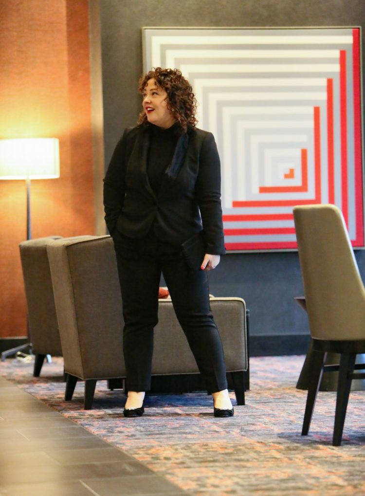 Alison in a black crepe tuxedo pantsuit with a black cashmere turtleneck walking through a hotel lobby
