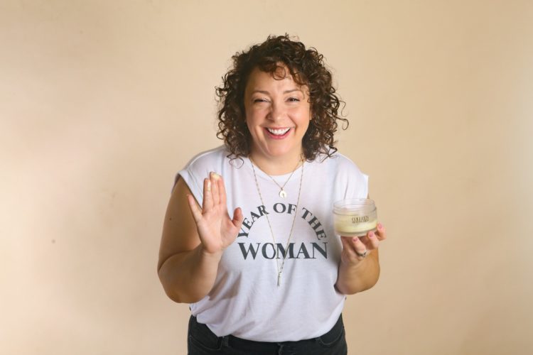 woman in a white t-shirt with an open jar of Colleen Rothschild Radiant cleansing balm, some of the balm on her finger as she smiles at the camera