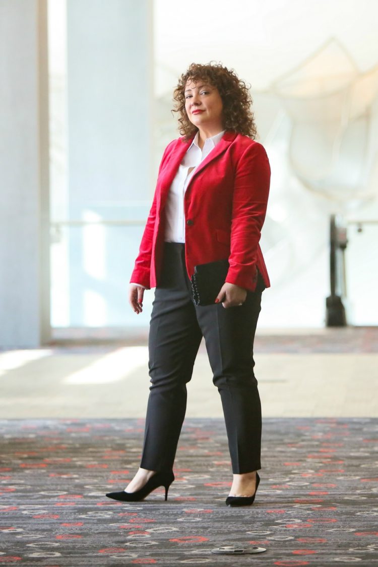 Alison in a red velvet blazer styled with a white button front shirt and black cigarette pants looking directly into the camera