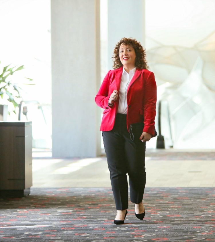 Alison in a red velvet blazer over a white shirt styled with black tux pants walking through a hotel lobby