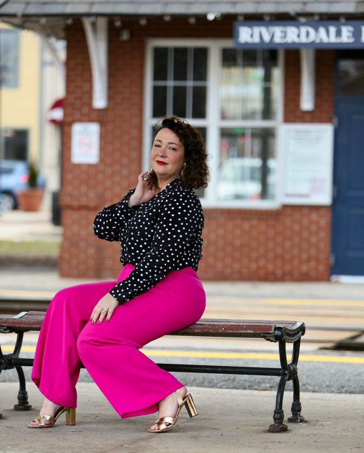 woman in pink pants and a black polka dot blouse sitting on a bench looking at the camera