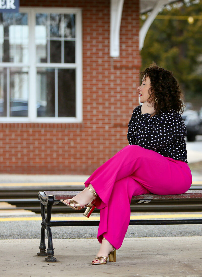 Woman in a polka dot blouse and hot pink pants with gold sandals sitting on a bench looking off into the distance