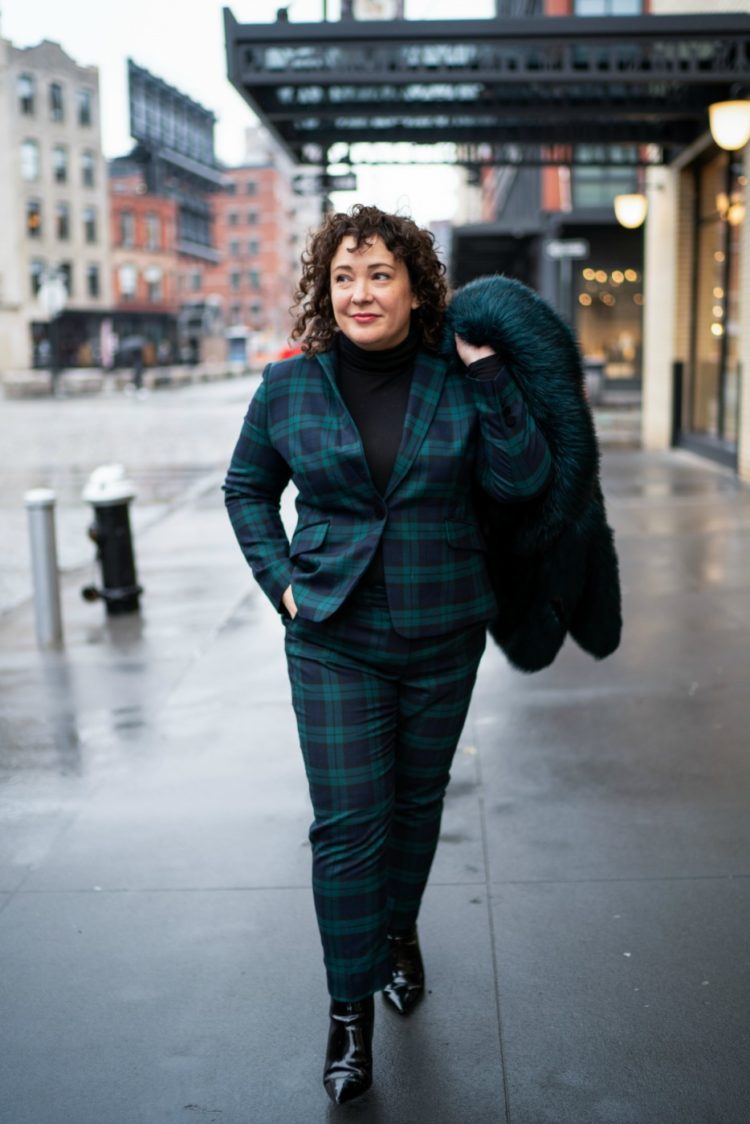 woman in a black watch plaid pantsuit carrying a dark green faux fur coat over her shoulder on the streets of New York City