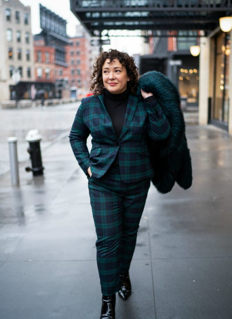 woman in a black watch plaid pantsuit carrying a dark green faux fur coat over her shoulder on the streets of New York City