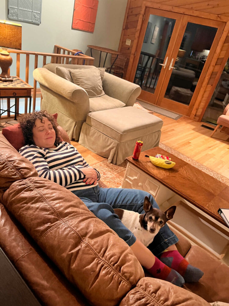 a woman in the living room of a lake house, lying on a sofa. She is wearing a navy and white striped shirt and jeans. Her eyes are closed, her arms crossed over her stomach. She is lying on her back and a toy fox terrier dog is sitting between her knees looking up at the camera