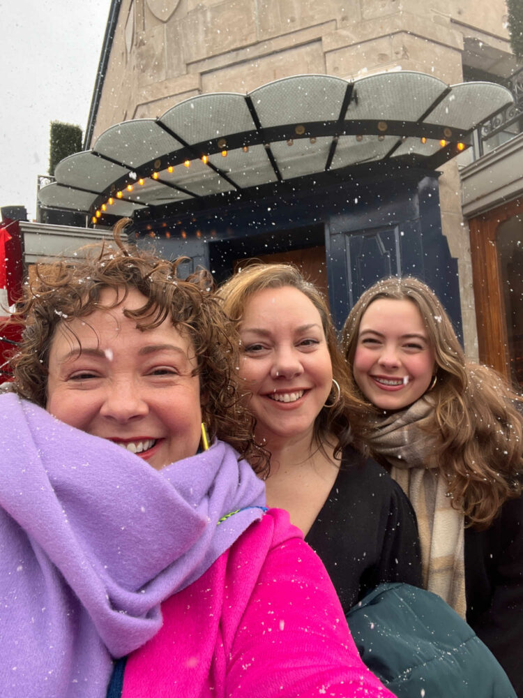 three women standing together in front of the Le Diplomate restaurant in Washington, D.C. It is snowing and they are smiling.