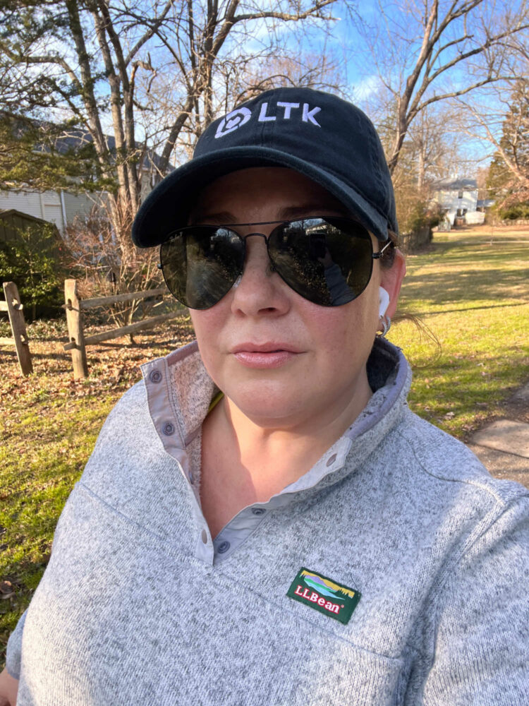 woman in a gray swater fleece pullover from L.L.Bean and a black baseball cap with the LTK logo on it. She is wearing black oversized aviator sunglasses from Ray-Ban and AirPods in her ears. She is outside on a walking path 
