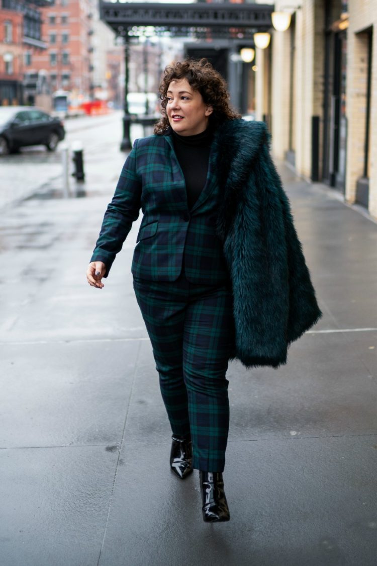 woman in a black watch plaid pantsuit carrying a dark green faux fur coat over her shoulder on the streets of New York City