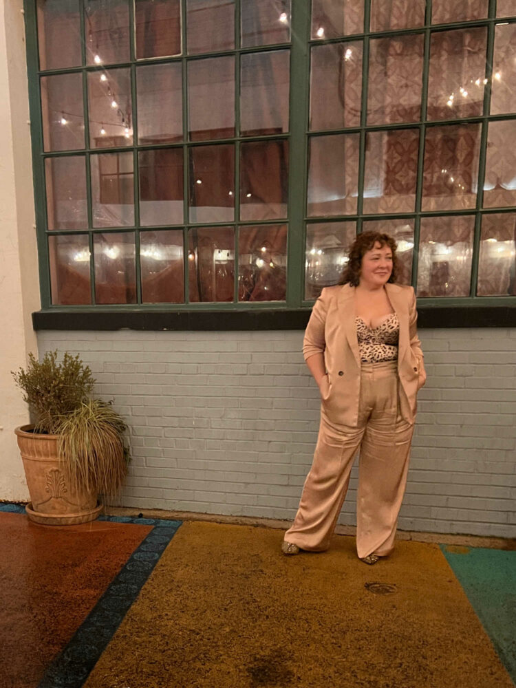 woman in a slouchy tan satin pantsuit with a tan leopard print satin corset underneath. She is standing in front of a building with old paneled windows, her hands are in her pants pockets and she is looking away from the camera