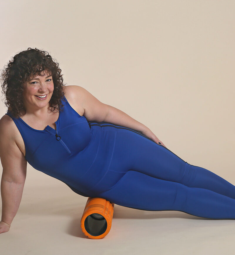 woman in blue bodysuit massaging her hip on a foam roller
