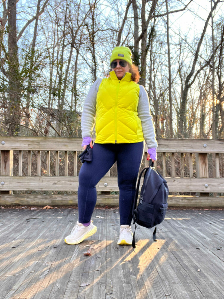 woman in a yellow vest, gray sweater fleece, navy thermal leggings, and white sneakers. She is holding a black backpack in her hand and is standing on a wood bridge
