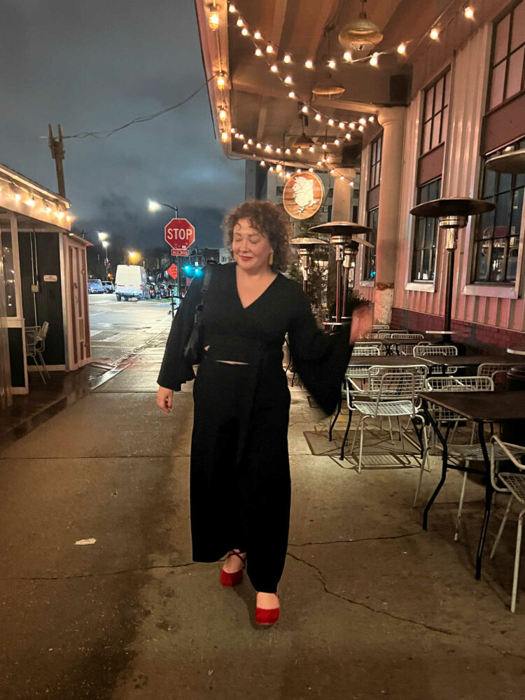 woman with curly brown hair wearing a black cropped wrap top with bell sleeves and black high waisted cropped wide leg trousers. She has styled the look with a Balenciaga city bag and red suede cross-strap platform heels