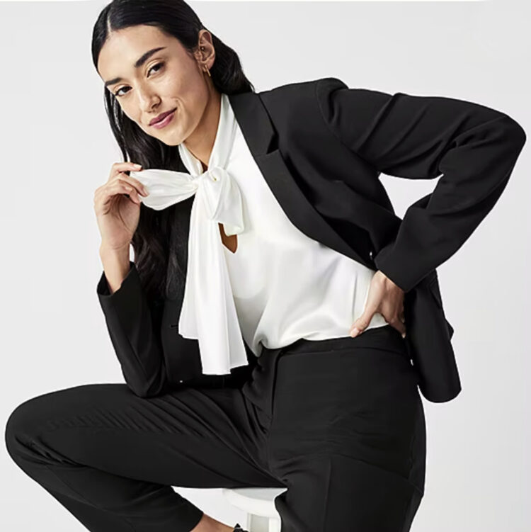 the best budget friendly petite suits for women are from JCPenney. This photo shows a black pantsuit and a white bow blouse from the brand Black Label by Evan Picone on a model who has long black hair parted in the middle. 