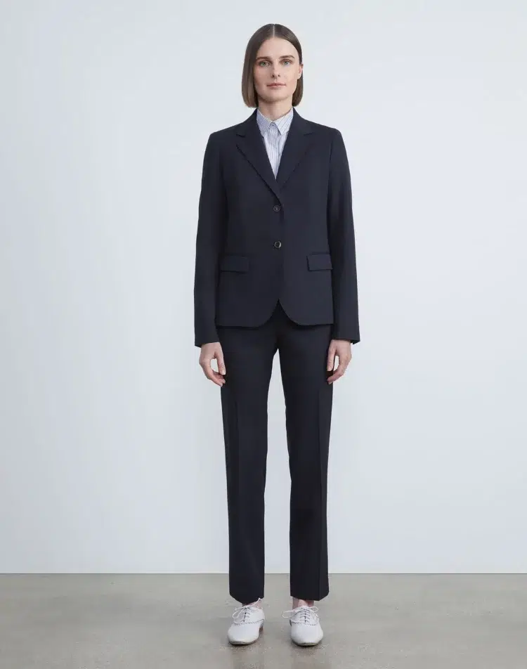 Lafayette 140 model with chin-length hair is wearing a navy blazer and matching cropped pants over a gray-blue silk button-up blouse and white oxford shoes on her feet. She is standing upright in front of a pale gray wall