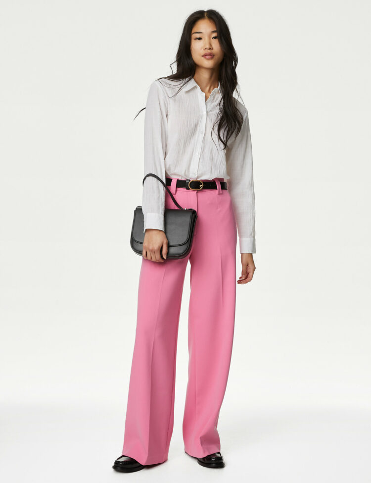 Asian woman in her 20's with long straight black hair to her shoulderblades. She is wearing a white blouse tucked into pink wide leg crepe trousers from M&S. She has them cinchd with a black belt, is wearing black loafers and holding a black leather clutch purse in her hand. She is standing on a white backdrop staring at the camera.