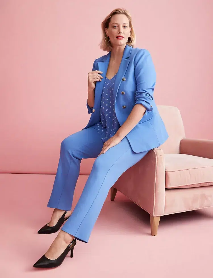 a Lane Bryant model with short blonde hair wearing a cornflower blue suit, sitting on the edge of a blush pink chair, holding the lapel of her blazer and looking at the camera