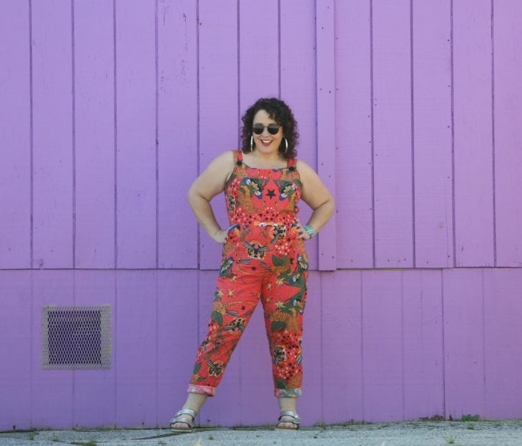 woman with dark brown shoulder length curly hair standing in front of a purple wall. She is wearing cotton overalls that fit more like a jumpsuit. They are an orange print with tigers all over them. She has styled the overalls with silver hoops, silver Birkenstock Arizona sandals, and a silver and turquoise cuff bracelet.