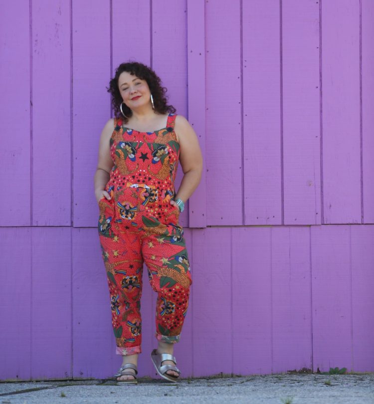 woman with dark brown shoulder length curly hair standing in front of a purple wall. She is wearing cotton overalls that fit more like a jumpsuit. They are an orange print with tigers all over them. She has styled the overalls with silver hoops, silver Birkenstock Arizona sandals, and a silver and turquoise cuff bracelet.