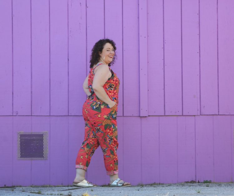 woman with dark brown shoulder length curly hair standing in front of a purple wall. She is wearing cotton overalls that fit more like a jumpsuit. They are an orange print with tigers all over them. She has styled the overalls with silver hoops, silver Birkenstock Arizona sandals, and a silver and turquoise cuff bracelet.