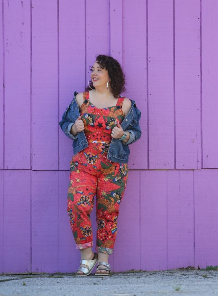 woman with dark brown shoulder length curly hair standing in front of a purple wall. She is wearing cotton overalls that fit more like a jumpsuit. They are an orange print with tigers all over them. She has styled the overalls with silver hoops, silver Birkenstock Arizona sandals, and a silver and turquoise cuff bracelet.