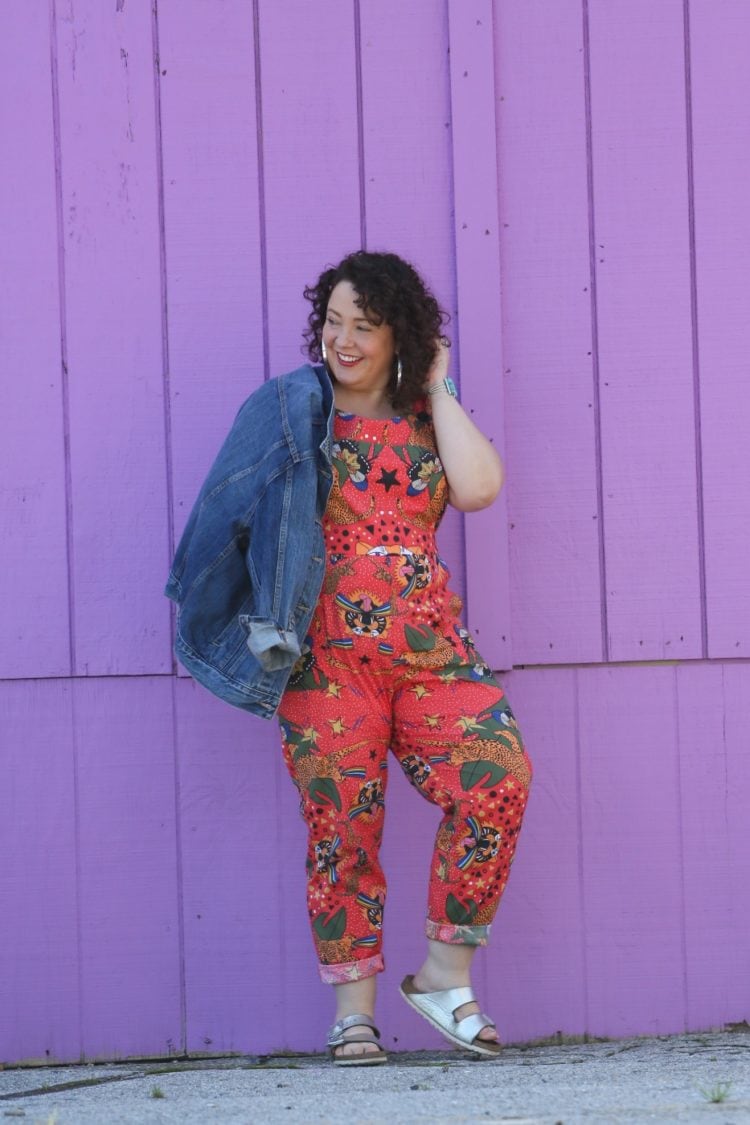 woman with dark brown shoulder length curly hair standing in front of a purple wall. She is wearing cotton overalls that fit more like a jumpsuit. They are an orange print with tigers all over them. She has styled the overalls with silver hoops, silver Birkenstock Arizona sandals, and a silver and turquoise cuff bracelet.