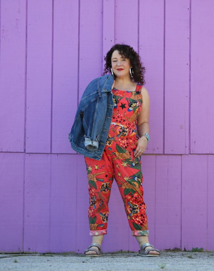 woman with dark brown shoulder length curly hair standing in front of a purple wall. She is wearing cotton overalls that fit more like a jumpsuit. They are an orange print with tigers all over them. She has styled the overalls with silver hoops, silver Birkenstock Arizona sandals, and a silver and turquoise cuff bracelet.