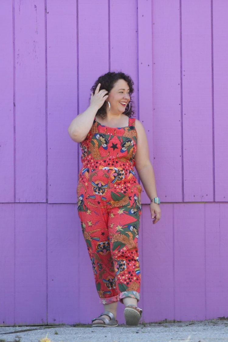 woman with dark brown shoulder length curly hair standing in front of a purple wall. She is wearing cotton overalls that fit more like a jumpsuit. They are an orange print with tigers all over them. She has styled the overalls with silver hoops, silver Birkenstock Arizona sandals, and a silver and turquoise cuff bracelet.