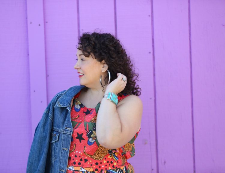 woman with dark brown shoulder length curly hair standing in front of a purple wall. She is wearing cotton overalls that fit more like a jumpsuit. They are an orange print with tigers all over them. She has styled the overalls with silver hoops, silver Birkenstock Arizona sandals, and a silver and turquoise cuff bracelet.