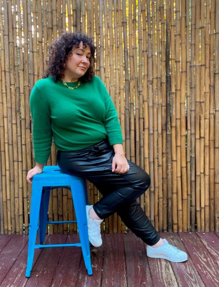 woman with brown curly hair sitting on a cerulean blue stool, wearing a green cashmere crewneck and black faux leather leggings