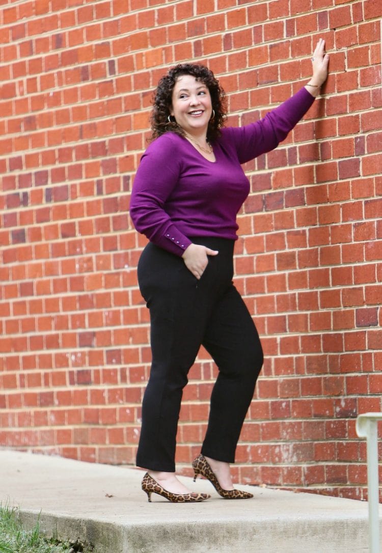 woman in black ponte knit work pants and a purple sweater and leopard kitten heel pumps leaning against a red brick wall