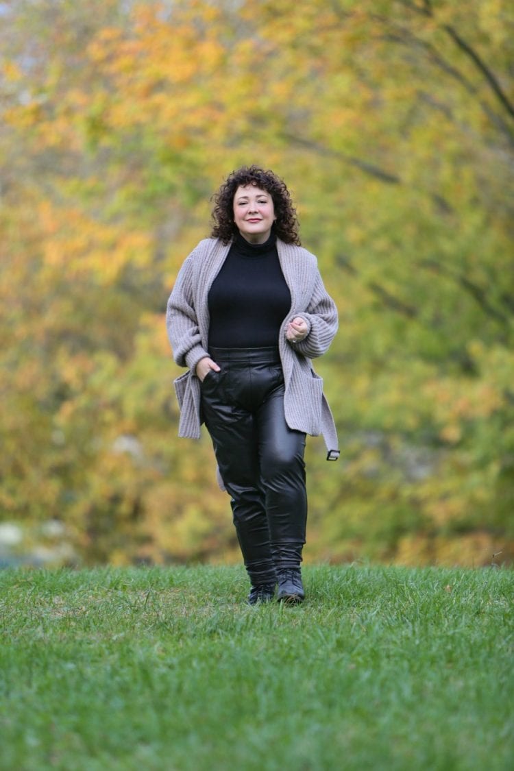 woman walking on grass in a field wearing a black turtleneck, taupe sweater coat, and black faux leather joggers from Spanx