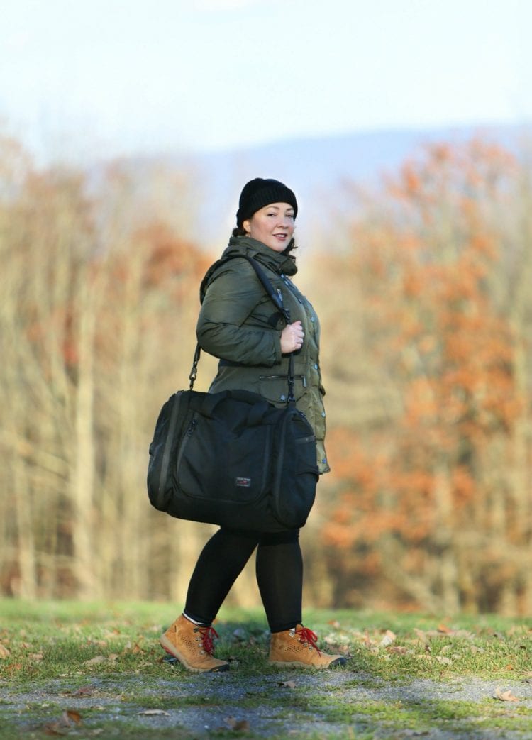 tom bihn aeronaut 45 rbag on the shoulder of a woman in a green anorak and black beanie, walking in the woods in autumn