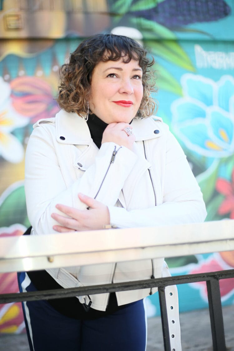 Alison wearing a white leather moto jacket. She is leaning against a railing with her hand resting on her fist, looking away from the camera.