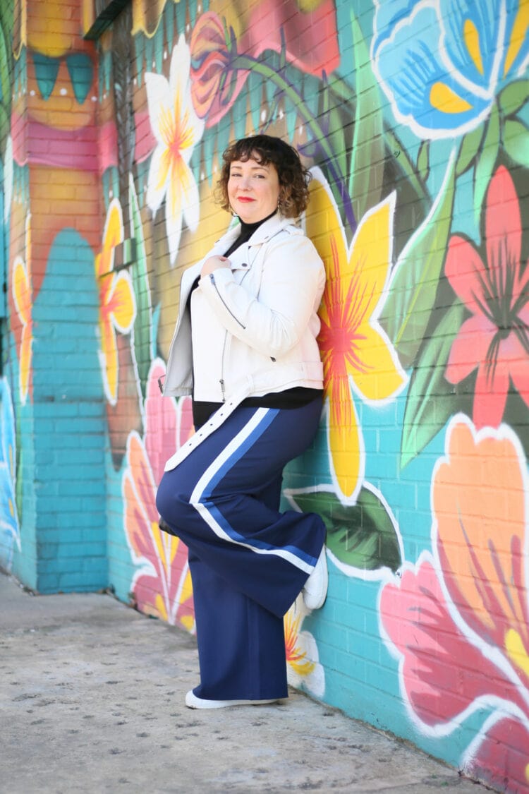Alison in the Universal Standard leeron moto jacket in white and the Stephanie pants in navy with blue and white racing stripes down the side. She is leaning against a mural with one foot up, holding the collar of her jacket and smiling at the camera.