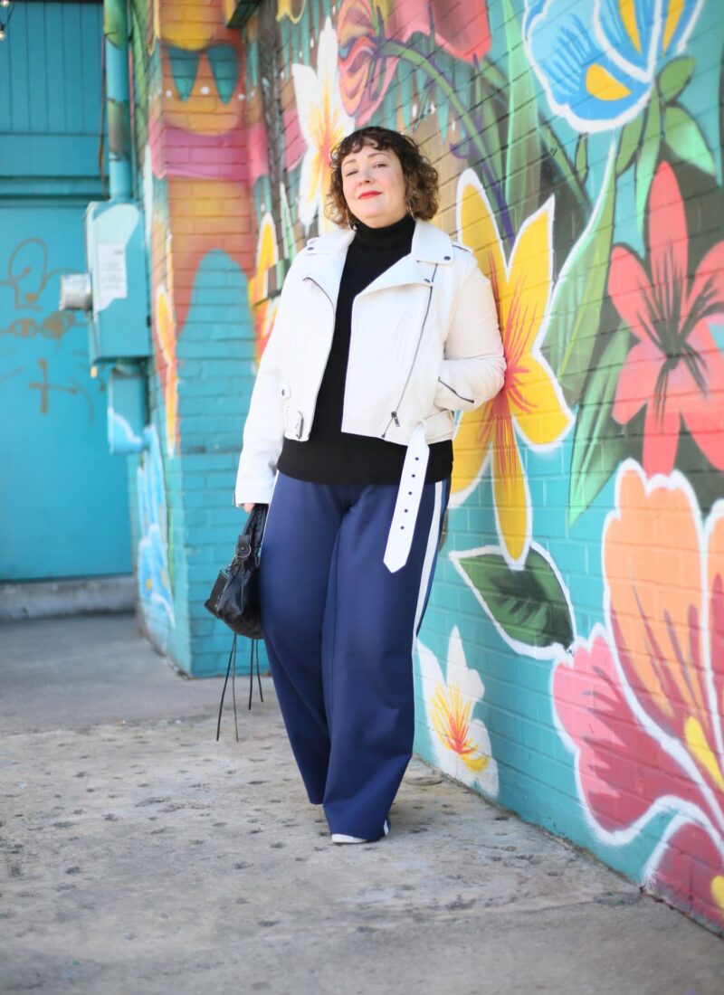 Alison leaning against a brick wall painted with a mural of tropical flowers. She is wearing a white leather moto jacket over a black turtleneck sweater with navy ponte wide leg pants and white sneakers. One hand is in her pocket and the other is holding a black leather Balenciaga City Bag.